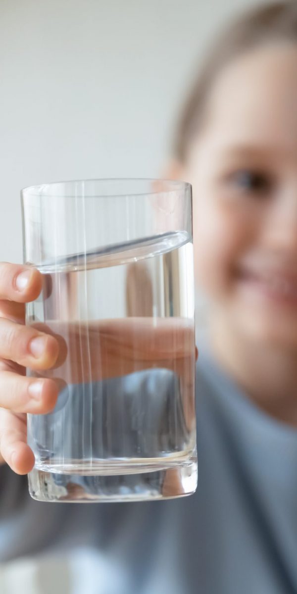 Close up smiling little girl holding glass of pure mineral water, offering to camera, cute pretty child kid recommending healthy lifestyle habit, drinking clean aqua for refreshment